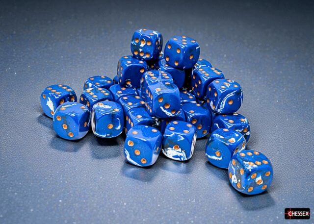 Blue dice with orange pips on a gray surface, brand 'Chessex' visible.