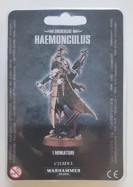 Packaged miniature figure of a Drukhari Haemonculus with a gun and surgical tools, from the Warhammer 40,000 series.
