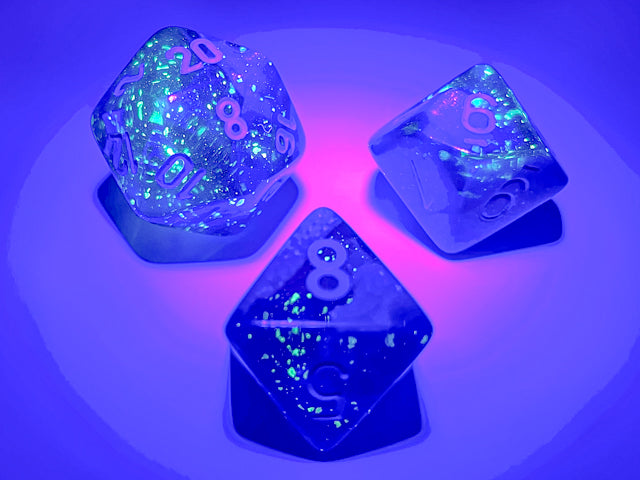 A close up example of some of the Gemini Blue-Blue with light blue polyhedral dice set under a blacklight to illustrate their luminescent features. 
