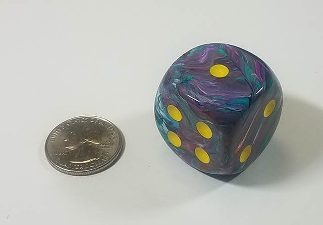 A multicolored dice with yellow spots next to a quarter for size comparison.