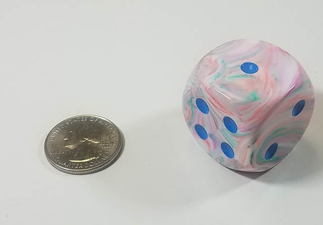 A swirled pink, white, and blue dice with blue spots next to a quarter for size comparison.