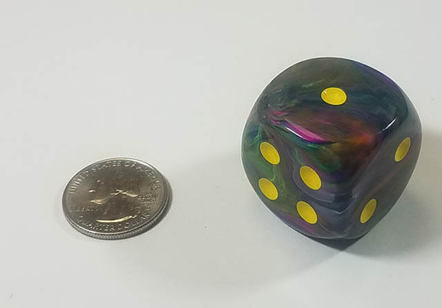 A multicolored dice with yellow spots next to a quarter for size comparison.