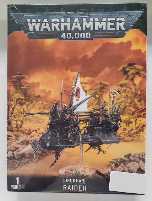 A product image featuring the Warhammer 40,000 Drukhari Raider box set, containing a multi-part plastic Raider with a large flying base, depicted in a dramatic sky backdrop.
