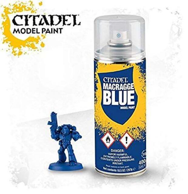 A can of Citadel model paint labeled 'Macragge Blue' with a spray nozzle on top. Next to it is a miniature figurine partially painted in blue.